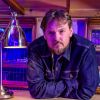 Close up profile picture of SW Hammond. He's looking into the camera, wearing a blue denim jacket, lit with dramatic shadows, and cool blue and purple lights seeping from the background.